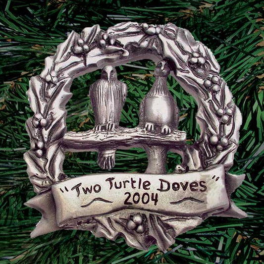 A pair of intricately detailed doves face forward on a shared perch in a wreath of holly bearing a banner with the words “Two Turtle Doves — 2004”. Hand carved then die-cast in pewter (palm sized, round, single-sided)