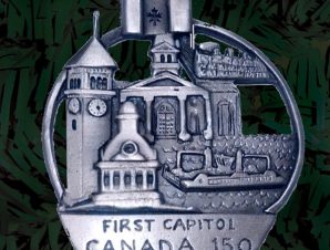 Finely detailed collage of sights in Kingston, Canada’s first capital: the Wolfe Islander III ferry, city hall dome, block tower, courthouse and Canada flag. Words read: First Capitol Canada 150 (palm sized, single-sided roundish, die-cast pewter)