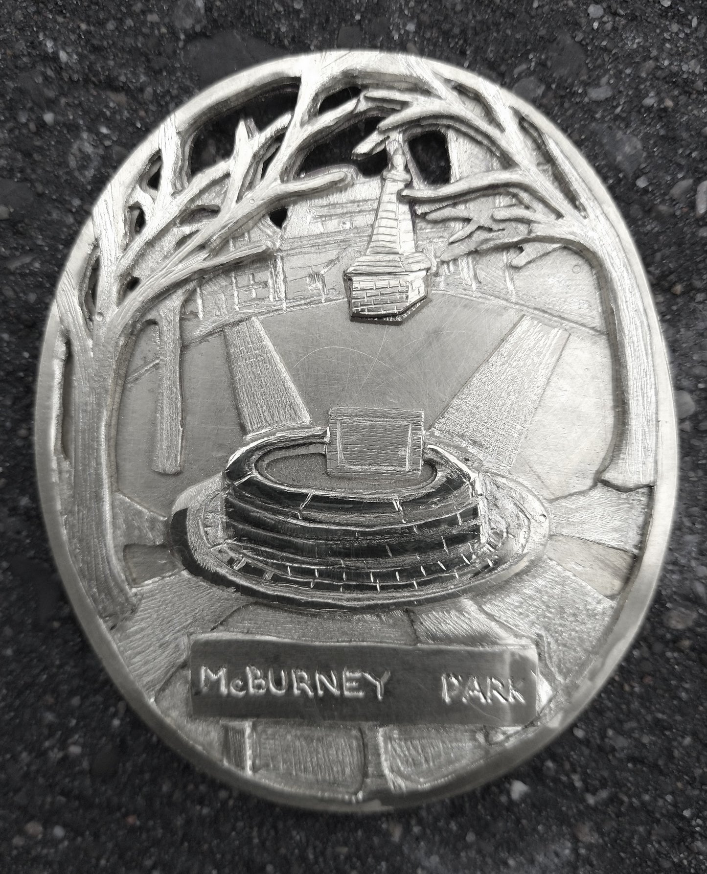 Finely detailed perspective of McBurney (Skeleton) Park in Kingston, Ontario, honed in on the central planter with backdrop of war memorial, bare trees, and neighbourhood houses (palm sized, single-sided oval, die-cast pewter)